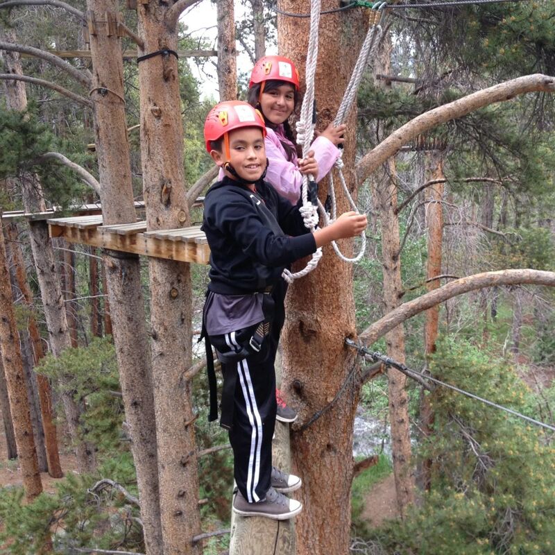 Two children wearing helmets are on a ropes course in a forest. The child in the foreground is wearing a black jacket and black and white striped pants, while the child behind is wearing a pink shirt. They are both holding onto ropes and appear to be enjoying the activity.